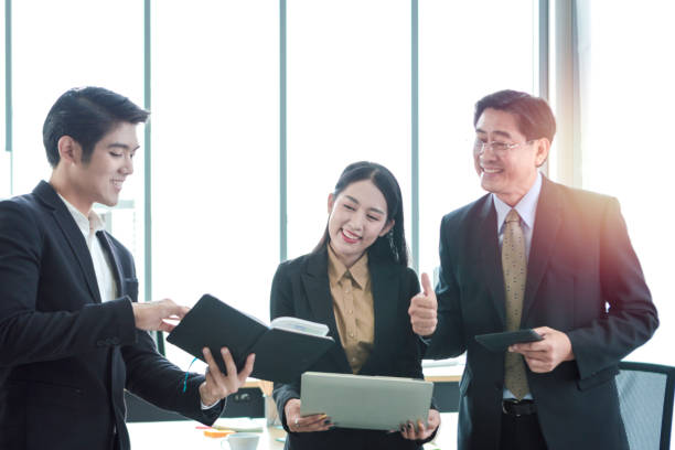 istock Happy business people standing and discussing about project work together in office, businesswoman holding laptop computer while young man was taking short note and sharing his idea on his notebook, senior businessman giving thumb up, people work together 1225587958
