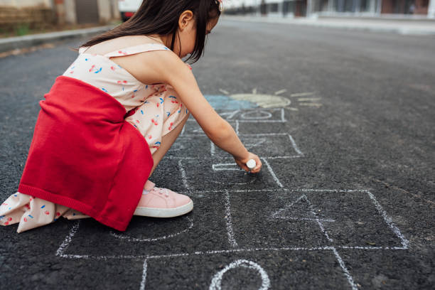 vista lateral vista al aire libre de dibujo de niña con tiza hopscotch en el patio de recreo. niño jugando el juego afuera. niño vestido durante el juego de lúpulo dibujado en el pavimento. actividades para niños - little girls sidewalk child chalk fotografías e imágenes de stock