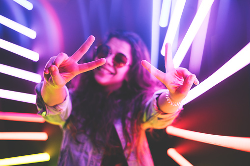 Cheerful cute smiling girl shows peace sign in round glasses at purple neon background.