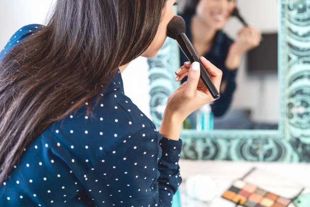Young woman applying makeup while looking in mirror - Happy girl using make up brush - Cosmetic and lifestyle people concept