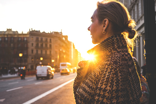 An adult beautiful lady in a plaid coat in the rays of the setting sun stands along the street of barcelona and looks away.  View assembly close.