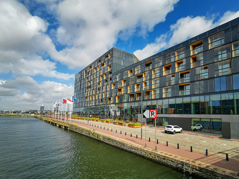 Amsterdam, Netherlands - August 11, 2019: Wide promenade runs along the bank of the IJ river. The walking passage tiled with red bricks and the car parking lot at the Hotel Jakarta Amsterdam laid with white floor bricks. Glass facade of the hotel is in the background.