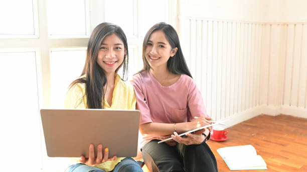 istock Photo of young sisters using a computer tablet and laptop while sitting together at the comfortable sitting room ground. Women with online shopping and relaxing time concept. 1225598379