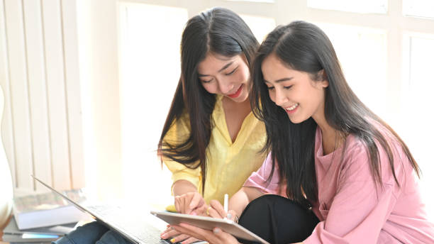 istock Photo of young sisters using a computer tablet and laptop while sitting together at the comfortable sitting room ground. Women with online shopping and relaxing time concept. 1225598505