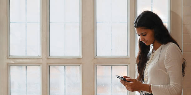 istock Beautiful French woman holding and using a smartphone in her hands while standing over comfortable living room windows as background. 1225598515