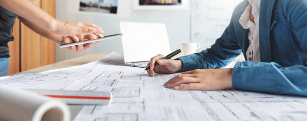 Asian Interior Designer and partner brainstorm and design in building Architecture project. Architect team consult and work for construction on digital tablet, laptop, paperwork, blueprint at office
