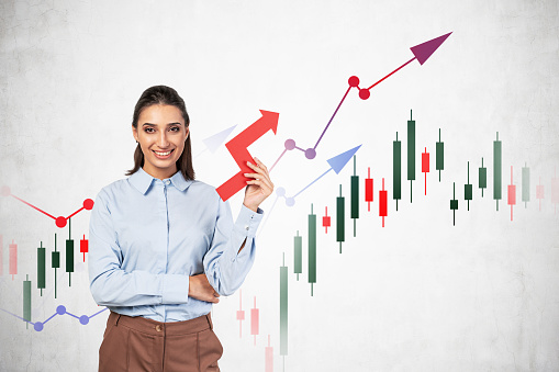 Cheerful young businesswoman with dark hair holding growing graph near concrete wall with financial charts. Concept of growth, success and investment