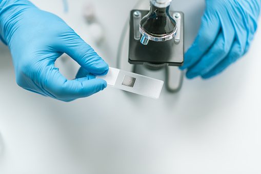 Overhead shot of a scientist wearing gloves inserting a slide into a microscope for a closer and more detailed examination.