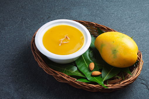 Aamras is plain Haapus or Alphonso Mango Puree/Pulp with almond topping. Aam Ras is a delicious Indian seasonal Dessert recipe. Served in a white bowl with mangos and almond placed in wooden basket on dark background
