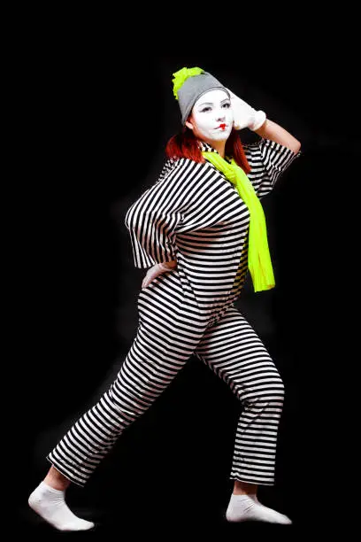 Portrait of female mime artist, isolated on black background. Young woman in striped suit and bright yellow scarf and hat is posing with her hand at her head.