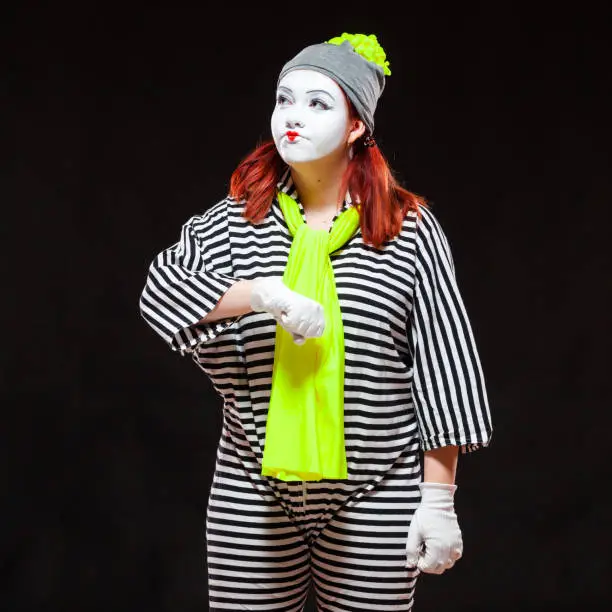 Portrait of female mime artist, isolated on black background. Young woman in striped clothes and bright yellow scarf is displeased with lateness looking at her watch. Symbol of waiting, time, punctuality, delay.