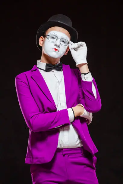 Portrait of male mime artist, isolated on black background. Young man in purple suit is touching his chin with a thoughtful look. Symbol of reflection, criticism.
