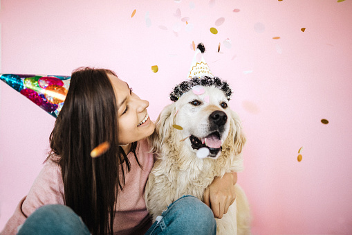 Golden retriever and cheerful young woman wearing party hats together, sitting on floor, confetti falling