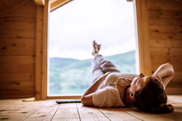 Rear view of woman lying on the floor in wooden house on the lake and listening music on headphones