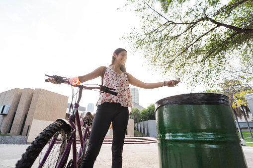 A happy young latin woman standing next to her bike in the city and throwing trash in a garbage can.