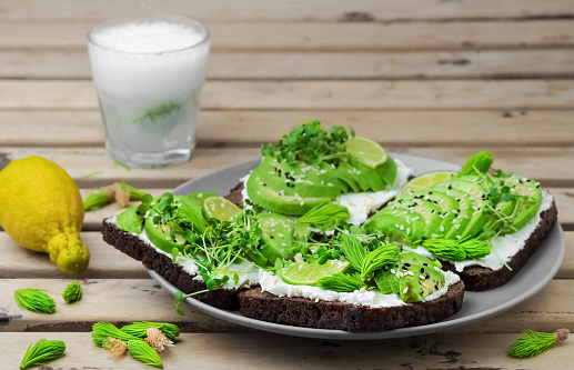 Sandwich on rye bread with avocado, cheese, microgreens, sesame seeds and spruce tips in a plate on a dark wooden table. Healthy vegetarian snack, closeup. Lemonade with lime on the table.