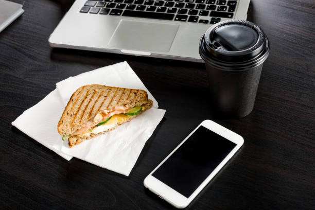 Coffee and a sandwich lying on the office desk next to the phone and laptop. A dark background. Business lunch concept during the work process.