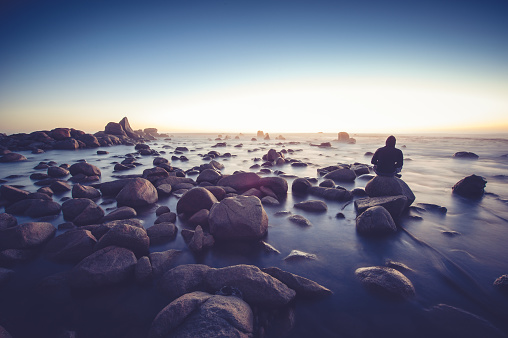 A lonely person sits on a rock at the ocean and stairs at the sunset. He is wearing a hoody. There is mist on the sunset horizon.