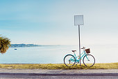 Vintage style bright bicycle parked near a blank road sign in the seafront quay. Sea view landscape with paving walking paths with a palm tree at sunset time. Calm and relax concept. Copy space.