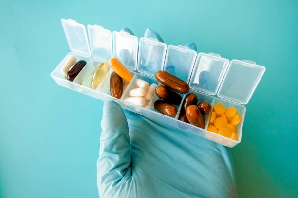 uma mão com uma luva médica contém cápsulas multicoloridas, uma caixa de comprimidos com medicamentos em um fundo azul - vitamin pill lecithin group of objects capsule - fotografias e filmes do acervo