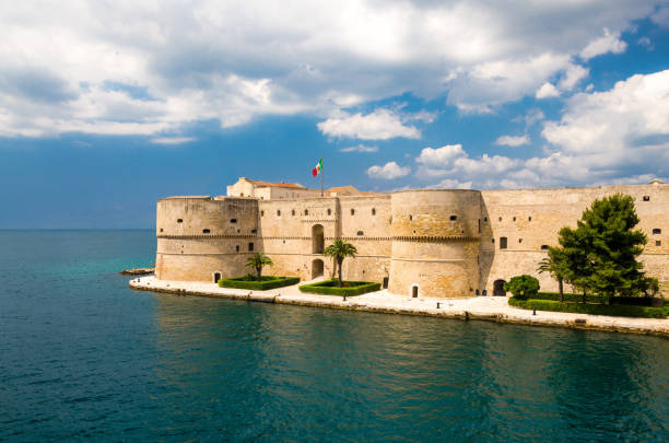 Old medieval Aragonese Castle, Taranto, Puglia, Italy Taranto , Italy, May 7, 2018: Old medieval Aragonese Castle with palm trees in front of blue sky with white clouds on sea channel in historic center of city, Puglia taranto stock pictures, royalty-free photos & images