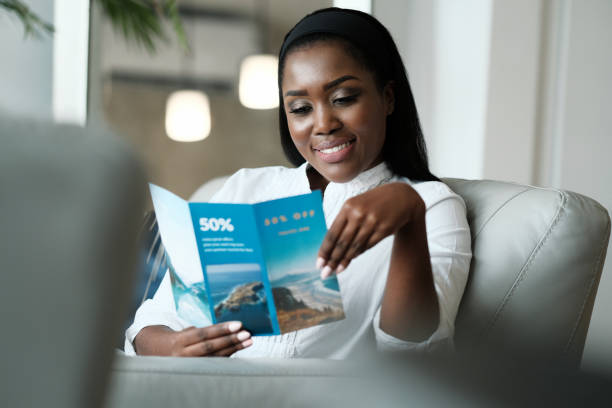 Black Woman Reading Travel Flyer For Holiday Trip African American Woman Sitting On Sofa, Reading A Travel Flyer Advertising Promotional Discounts For Vacations. pamphlet stock pictures, royalty-free photos & images