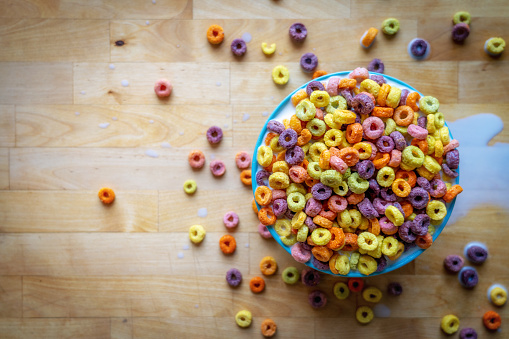 Bowl of colourful breakfast cereal spilling over wooden table top, flat lay.