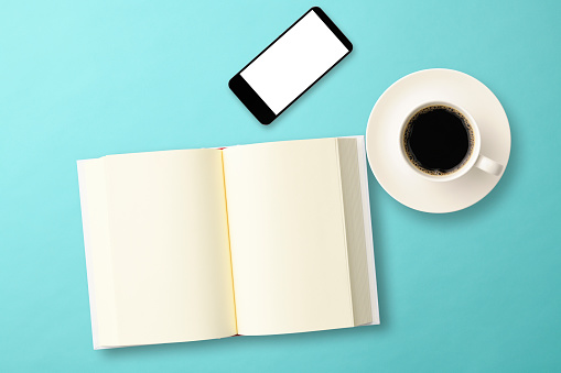 Overhead shot of blank book with coffee and smart phone on light blue background.