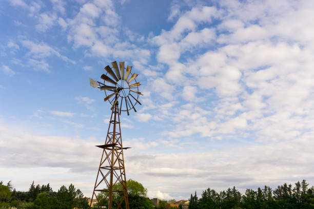 The old rusting steel windmill is a machine for producing continuous power from wind and air, in which a wheel fitted with steel vanes, trees in the background under white fluffy cloudy and blue sky