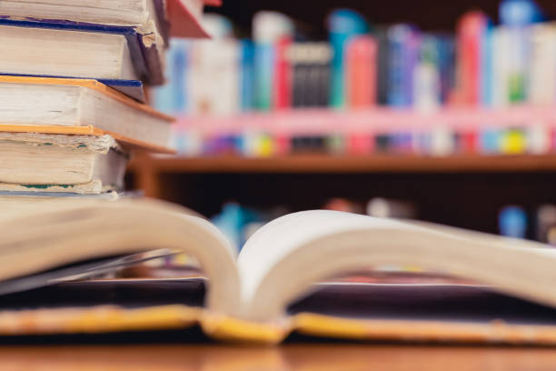 istock Textbook stack on wooden table with blur background of bookshelf and opened book in library. Photo concept of education, knowledge, traditional learning, and literature. Selective Focus 1225725673
