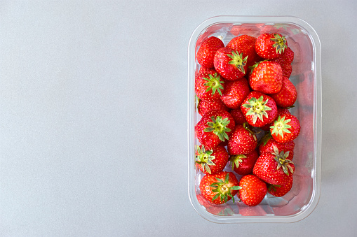 Fresh ripe strawberries in a clear plastic container on grey background. Flat lay image with copy space.