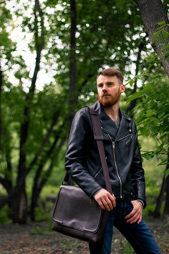 a man with a red beard and a bag over his shoulder in a black leather jacket stands in the forest and looks thoughtfully into the distance.