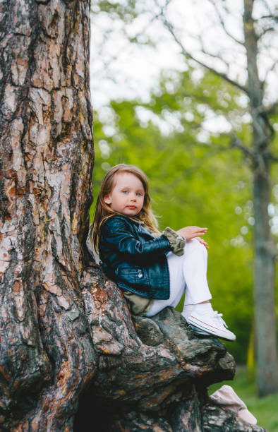 istock Little girl in leather jacket outdoors in a park sitting on tree trunk in spring day 1225740470