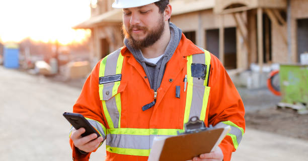 знамя строителя просмотра на смартфоне и проведение ноутбука - protective workwear hat violence construction стоковые фото и изображения