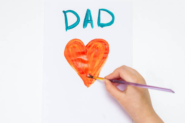 Top view of hand of child drawing red heart with word Dad greeting card by watercolors on white paper. Father's Day and family concept.