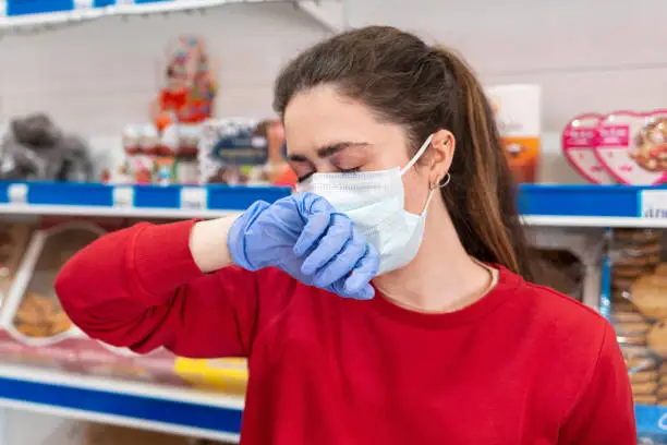 Photo of A woman in rubber gloves and a medical mask,suffering from a runny nose covering her face with her hand.In the background are shelves of the store.The concept of coronovirus and the crisis in business