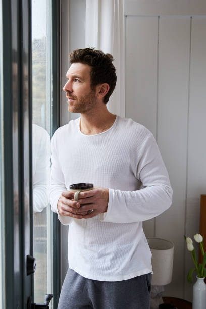 homme buvant le café tout en regardant par la fenêtre de sa chambre à coucher à la pluie - homme mug regarder dehors photos et images de collection