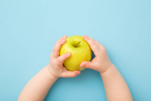 水色のテーブルの背景に緑のリンゴを保持している赤ちゃんの手。パステルカラー。新鮮なフルーツ。クローズ アップ。視点ショット。トップダウンビュー。 - child eating apple fruit ストックフォトと画像