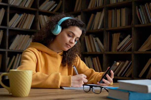 latin teen girl wear headphones hold phone online learning in mobile app. hispanic college student using smartphone watching video course, zoom calling making notes in workbook sit in library campus. - homework teenager mobile phone school imagens e fotografias de stock