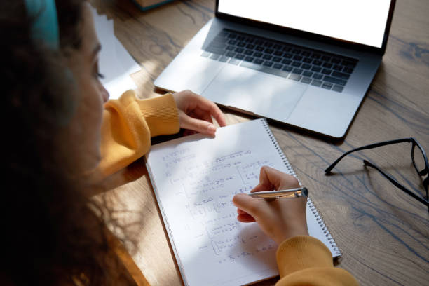 teen girl college student wear headphones studying from home writing in workbook solving equations learning math sits at desk. teenage school pupil learn online on laptop, close up over shoulder view. - homework imagens e fotografias de stock