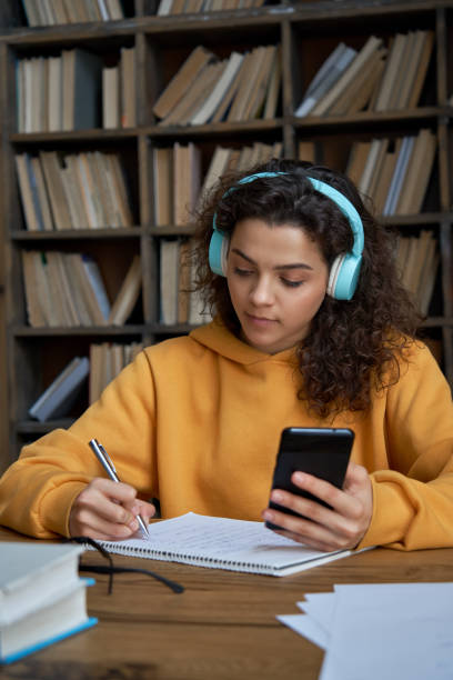 Hispanic teen girl school student wear headphones hold smartphone using distance learning mobile app online watching video course or zoom calling making notes in workbook sit at home or in library. Hispanic teen girl school student wear headphones hold smartphone using distance learning mobile app online watching video course or zoom calling making notes in workbook sit at home or in library. education student mobile phone university stock pictures, royalty-free photos & images
