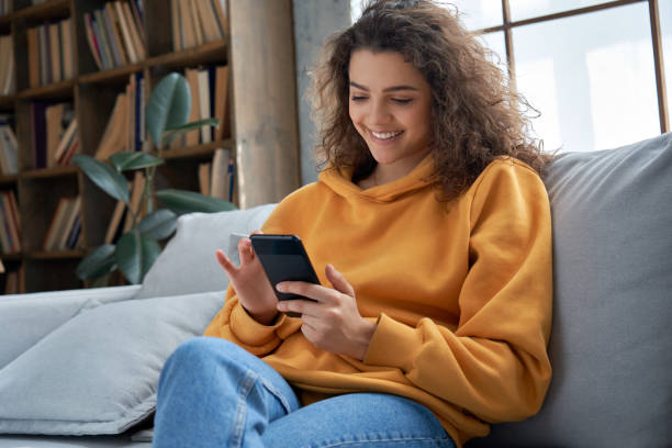 快樂千禧年西班牙少女檢查社交媒體持有智慧手機在家裡。微笑的年輕拉丁女人使用手機應用程式玩遊戲,網上購物,訂購送貨放鬆在沙發上。 - happy woman 個照片及圖片檔
