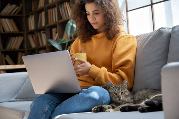 Young hispanic latin ethnic teen girl relaxing sit on comfortable sofa with cute pet cat watching remote education webinar class, movie series on laptop drinking warm tea in cozy sunny living room. joven hispano latino étnico adolescente adolescente relajante sentarse en cómodo sofá con lindo gato mascota viendo clase de webinar de educación remota, series de películas en el ordenador portátil beber té caliente en la acogedora sala de estar so