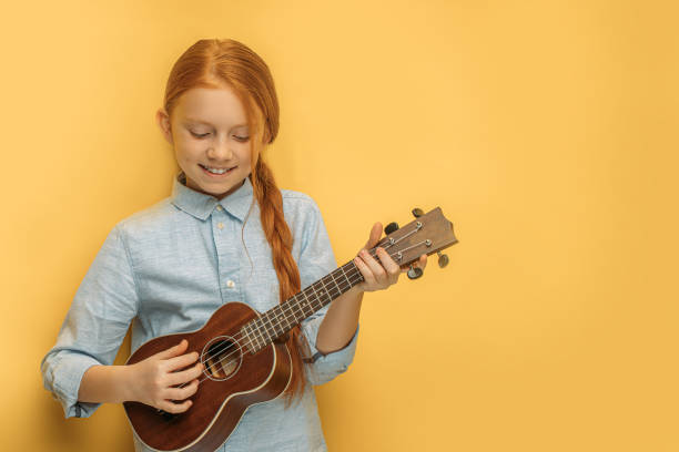 beautiful girl play ukulele isolated beautiful caucasian child girl playing ukulele, little girl with natural red hair and freckles loves music and instruments. isolated yellow background ukulele stock pictures, royalty-free photos & images