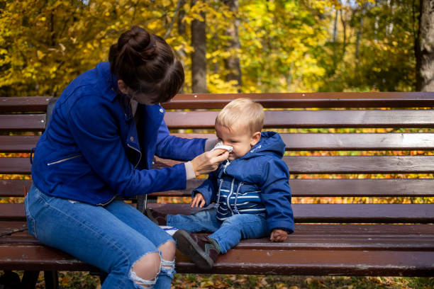 autumn cold in children - concept. a young mother in blue clothes on a park bench wipes the nose of little baby who has a runny nose and blows nose. close-up, soft focus, on background  trees in blur stock photo