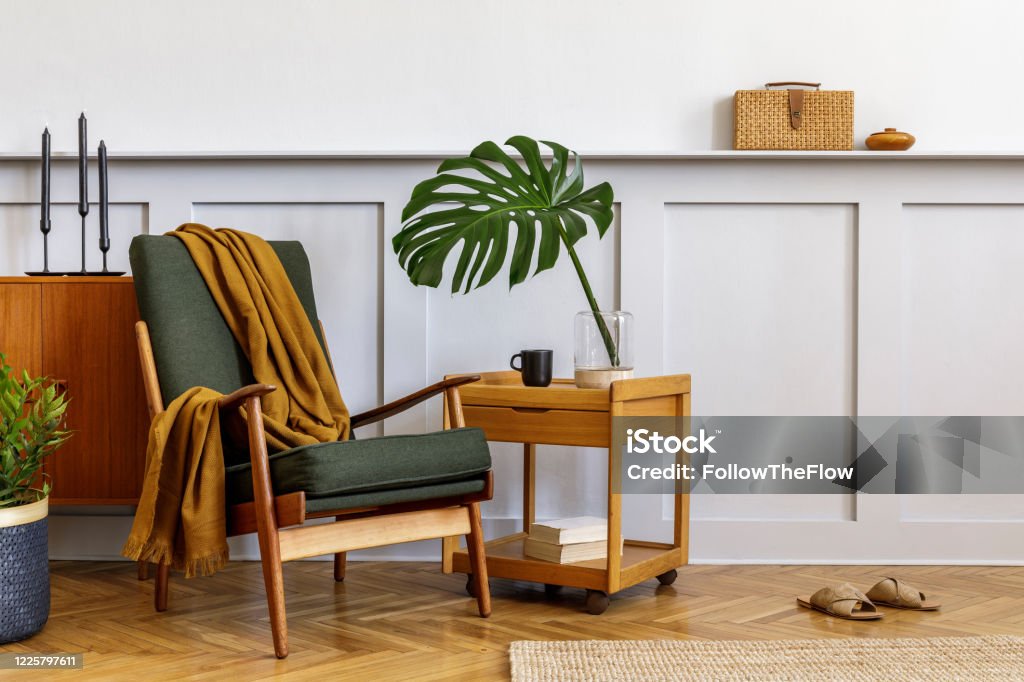 Interior design of stylish living room with vintage green armchair, wooden coffee table, furniture, grey wall, shelf, carpet, plants, leaves in vase, book, copy space and elegant personal accessories. Interior design of living room with design armchair, furniture and elegant personal accessories. Template. Home decor. Apartment Stock Photo