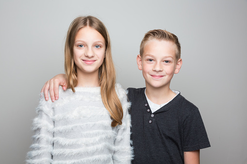 Double portrait in studio, in front of bright grey background: younger brother and teenage sister, looking into camera and smiling, boy with hands on girls shoulder