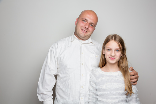 Double portrait in studio, in front of bright grey background: father and teenage daughter, standing and looking into camera, father with one hand on girls shoulder