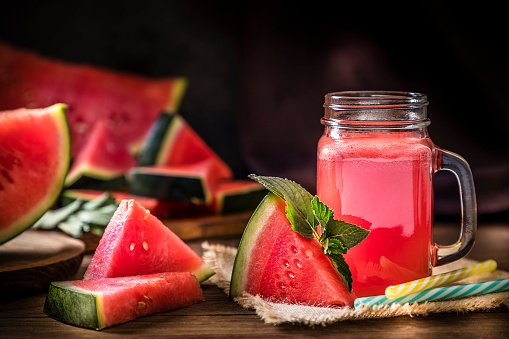 Front view of a dinking glass full of watermelon juice alongside a watermelon slice with mint leaves and two drinking straws. At the background is a defocused sliced watermelon. Low key DSLR photo taken with Canon EOS 6D Mark II and Canon EF 24-105 mm f/4L