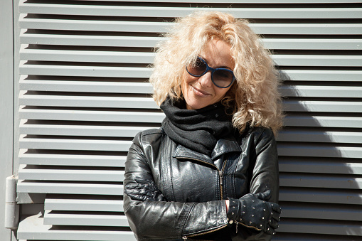 outdoors portrait of a 45 year old blonde woman with curly hair in a black leather jacket and sunglasses